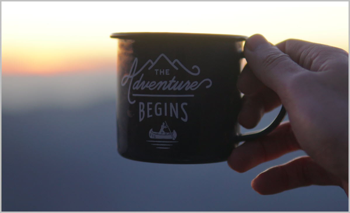 A hand holding a mug that reads The adventure begins, overlooking the mountains